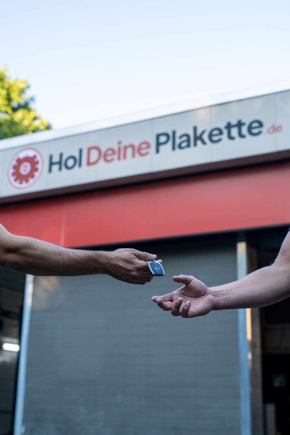 Zwei Arme sichtbar, wo eine Person die Autoschlüssel an eine andere übergibt, sie stehen vor einer Hauptuntersuchungsstelle